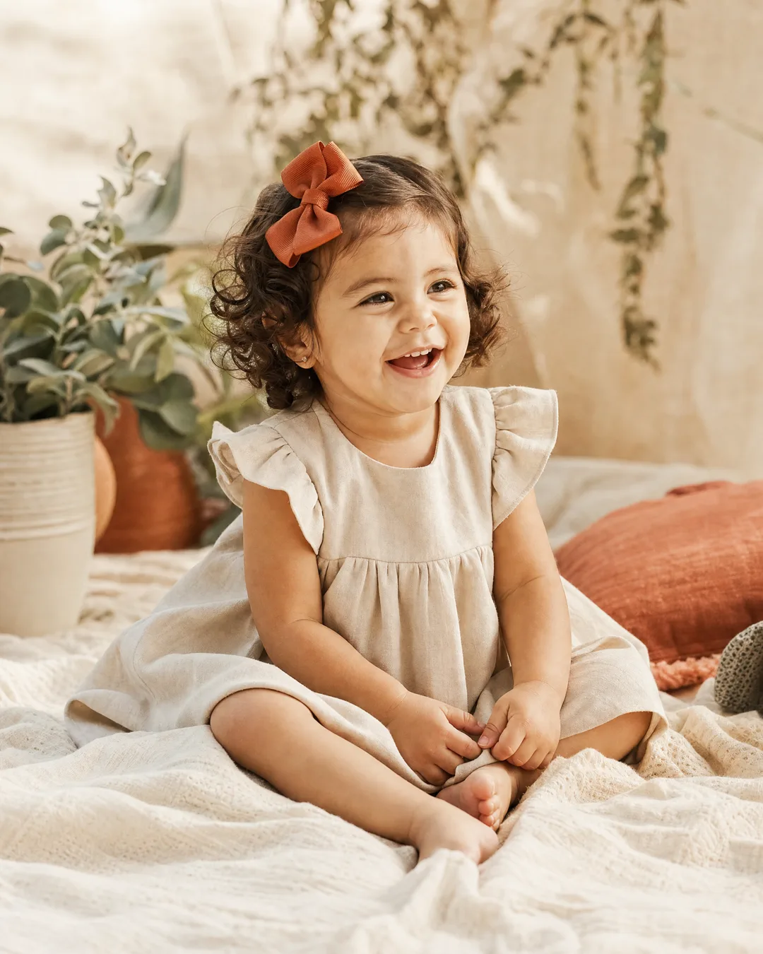Menina pequena sorrindo com laço terracota no cabelo, sentada sobre linho marfim
